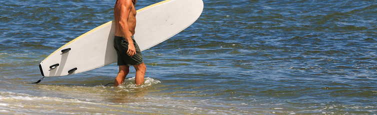 Gilgo Beach, New York, USA - 22 July: Side view of a male surfer walking into the ocean carrying his surfboard under his arm to go surfing on Long Island.