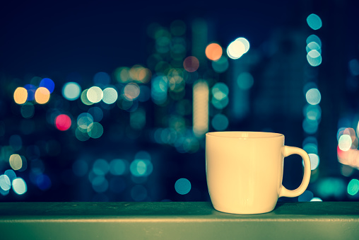 White cup over night cityscape light bokeh background - vintage tone. Drinking, relaxation time concept.