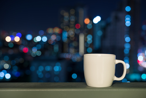 White cup over night cityscape light bokeh background. Drinking, relaxation time concept.