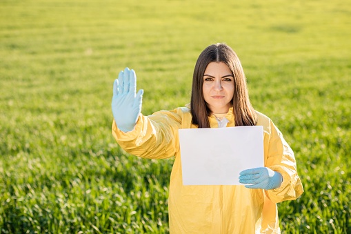 Person in overalls holds paper with blank sheet of paper for text inscription while standing on green field on sunset and the other hand shows a gesture to stop and not pollute the planet. Concept of bad ecology and environmental protection