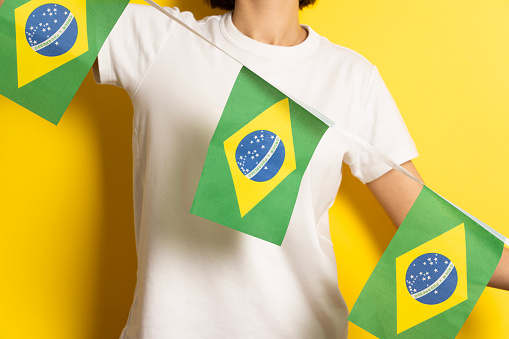A woman wearing a white t-shirt is standing against a yellow backdrop holding Brazil flags in her hands.