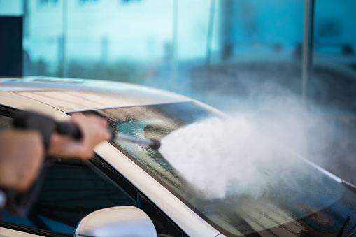 Man washes his gray car at a self-service car wash. Close-up of high-pressure water washing. A nozzle that sprays a stream of water on the windshield. Background: aesthetic car wash