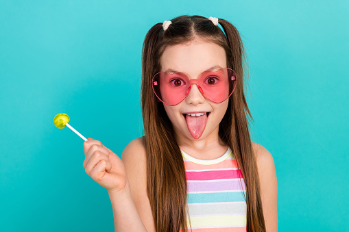 Portrait of excited cheerful schoolkid hand hold lollipop candy showing tongue out isolated on turquoise color background.