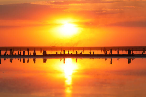 Orange sunset, dramatic and majestic sky over beach with sun reflection in water. Wood posts for salt extraction of extremely salty lake, Ukraine. Summer season, idyllic peaceful dusk sunlight.