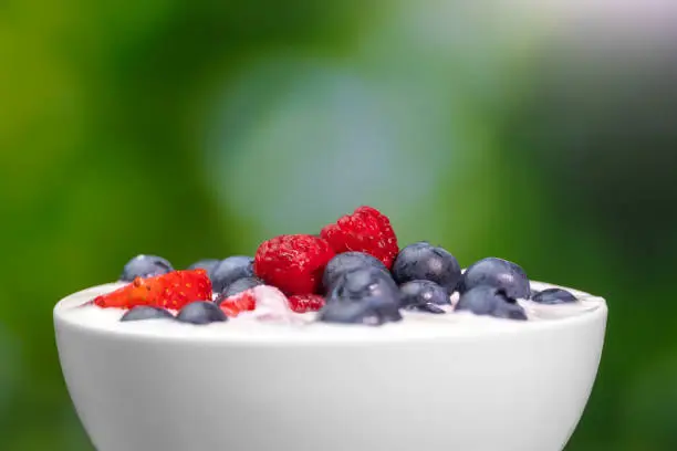 Photo of Close-up of a bowl with natural white yogurt and fresh raspberries, blueberries, strawberries on a green blurred background, macro photography. Delicious healthy breakfast