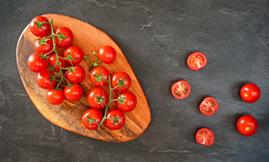 Fresh red cherry tomatoes on wooden cutting board, some sliced to half over black slate like board near