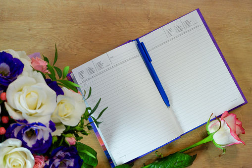 Notepad, diary with a pen on a wooden table near a bouquet of multi-colored roses. Workspace with diary and pink white flower on wooden background. Top view