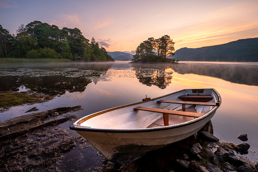 A wooden rowing boat on the shoreline of Derwentwater in The English Lake District. A beautiful sunrise can be seen in the sky with glassy reflections in the calm still water.