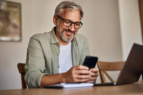 istock A smiling mature man typing on a mobile phone while working over a laptop at the home office. 1616719782