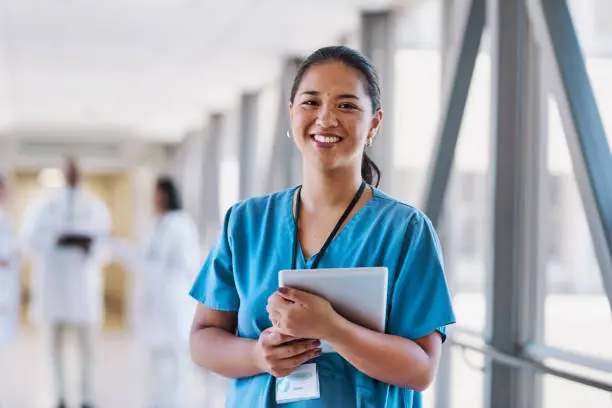 Female nurse carrying digital tablet smiling cheerfully at camera in hospital corridor, coworkers in background