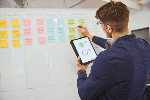 Businessman reading statistical chart and graph on digital tablet, standing in front of whiteboard and pointing at sticky note in office