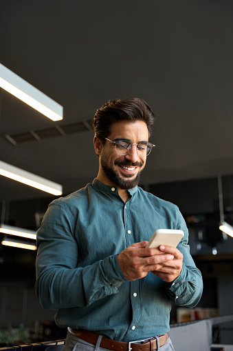 Smiling young Latin business man executive, businessman manager standing in office holding smartphone using apps on mobile cell phone managing digital apps on cellphone at work. Vertical