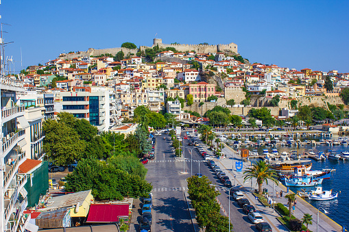 Kavala old town with its Byzantine castle in background, modern style houses by the street and harbour with fishing boats in foreground. Kavala town,  Makedonia, Greece.