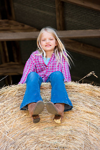 Midwestern Little Cowgirl Sits on Bale of Hay Barn Farm A midwestern little cowgirl, with plaid shirt and cowboy boots, sits on a hay bale in the storage barn on a farm. Americana. checkered cowboy boots stock pictures, royalty-free photos & images