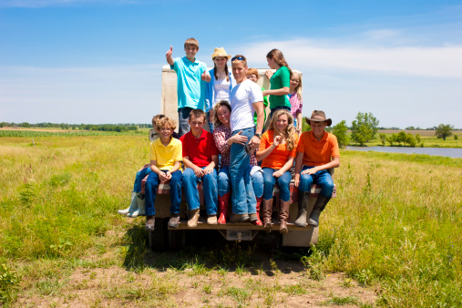 A proud hardworking midwestern family rides on the bed of a working farm  truck over the family farm. Vast expanse of open, fertile land spreads out beyond. Scene  represents 
