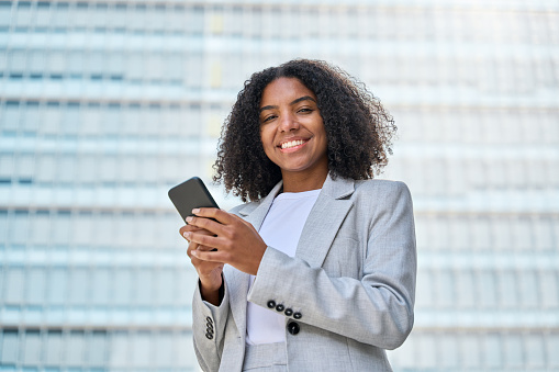 Young happy professional African American business woman wearing suit holding cell phone, looking at cellphone, using mobile apps on smartphone standing on city street, portrait.