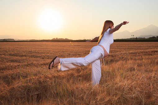 Young pregnant female enjoying the late sun. Flying pose. On one leg and arms in the air. Woman in white.