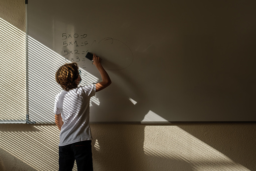 Back view of faceless schoolboy erasing answers of mathematical examples while standing near whiteboard in classroom during lesson
