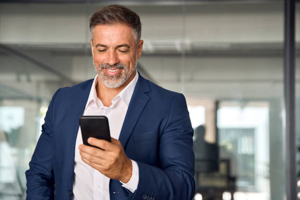 Smiling mature Latin or Indian businessman holding smartphone in office. Middle aged manager using cell phone mobile app. Digital technology application and solutions for business success development. stock photo