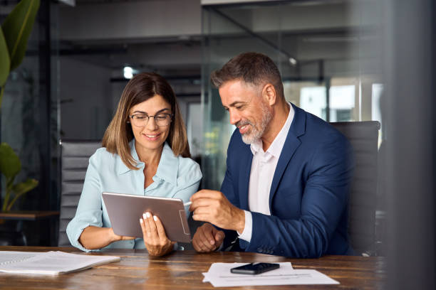 Team of diverse partners sitting at table mature Latin business man and European business woman discussing project on tablet in office. Two colleagues of professional business people working together. stock photo