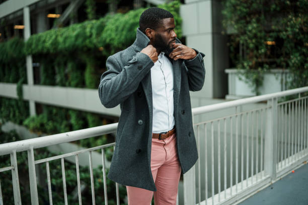 Young man of African American ethnicity wearing business casual clothing puts on a nice jacket while waiting outside on a bridge before walking into an airport.