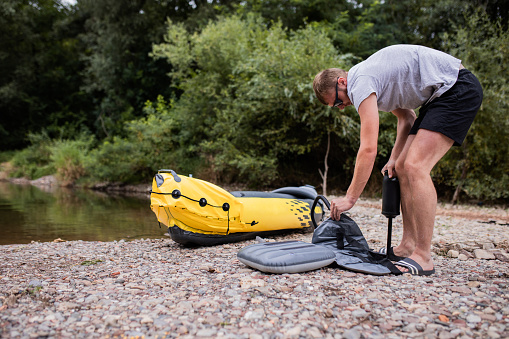 Young bearded man, an athlete, is inflating his rubber boat with a pump on the river shore. He is ready for kayaking.