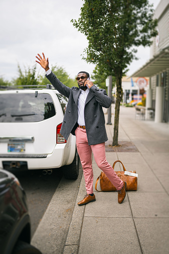 Well-dressed black businessman on a work trip stands on the sidewalk of a city street and hails a cap while taking a phone call.