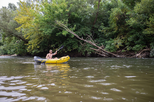 Young sportsman is rowing with an oar on the river in his yellow inflatable canoe. Wonderful nature is around him. Idyllic scene.