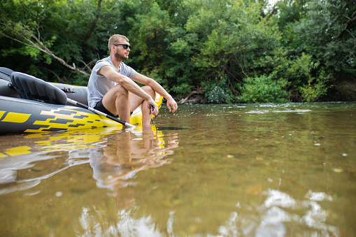 Young adventurous man, an athlete, with sunglasses is sitting with on his yellow inflatable canoe and looking at the river. Wonderful nature is around him. Idyllic scene.