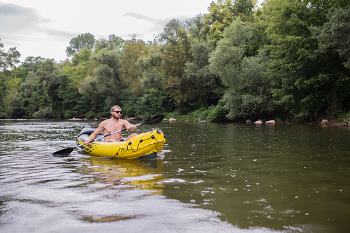 Young adventurous bearded man with sunglasses is rowing with an oar on the river in his yellow inflatable canoe. Wonderful nature is around him. Idyllic scene.