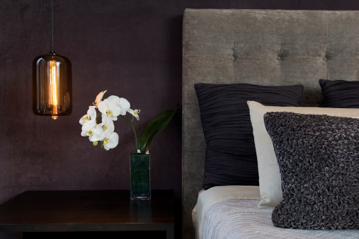 Gray headboard detail of a bed with textured pillows, lamp and white orchid flower.