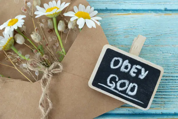 Obey God, handwritten text with chalk on small blackboard, vintage paper and flowers on wooden table. Top view. Christian biblical concept of obedience and faith Jesus Christ.