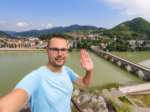 Young cheerful man having a video call on a smartphone while enjoying the view of Mehmed Pasa Sokolovic bridge (Na Drini cuprija) in Visegrad, Bosnia and Herzegovina.