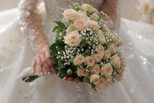 A young woman wearing a white wedding dress with an abundance of delicate flowers delicately holds a bouquet of beautiful white flowers
