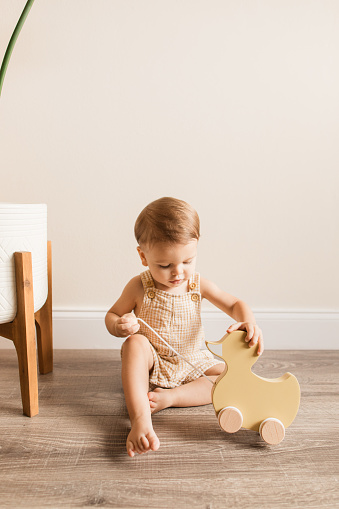 A Cute 20-Month-Old Baby Boy Wearing Organic Cotton, Yellow & White Plaid Overalls While Playing Barefoot on the Floor with a Wooden Rolling Yellow Ducky Toy at Home