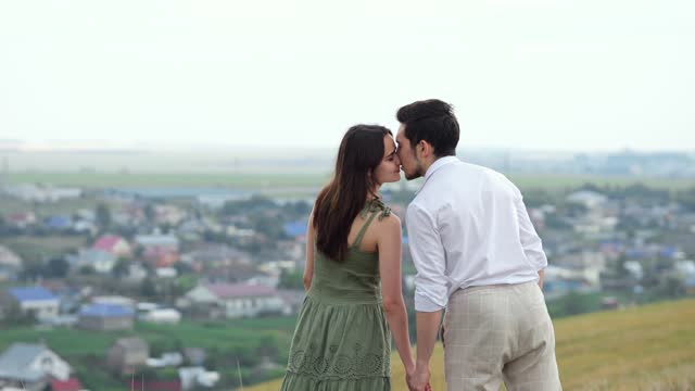 Young couple stands kissing on hill against village downward