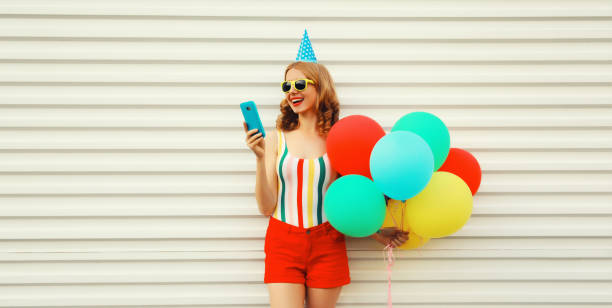 Happy cheerful young woman in festive birthday hat celebrating with smartphone and bunch of colorful balloons having fun wearing striped t-shirt, sunglasses on white background stock photo