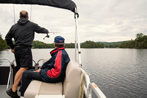 Senior son and elderly father enjoying fishing on a pontoon boat tour on a lake in summer. They are family, senior dad is in his eighty, son in his fifties. They are wearing casual clothes. Horizontal outdoors full length shot with copy space. This as taken in the Laurentides, Quebec, Canada.