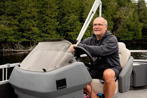Senior man driving his pontoon boat tour on a lake in summer, enjoying nature. He is in his fifties and is wearing casual clothes. Horizontal outdoors waist up shot with copy space. This as taken in the Laurentides, Quebec, Canada.