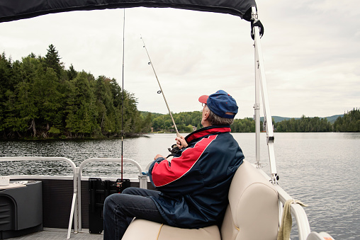 Elderly man enjoying fishing on a pontoon boat tour on a lake in summer. He is in his eighties and is wearing casual clothes. Horizontal outdoors waist up shot with copy space. This as taken in the Laurentides, Quebec, Canada.