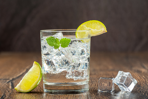 Cold refreshing sparkling water in a glass with ice, lemon or lime and mint. Wooden background and melting ice cubes. Copy space.