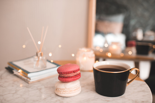 Cup of coffee with macarons and stack of paper books with candle on marble table in bedroom top view closeup.