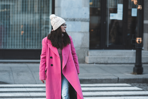 Happy stylish woman wear pink winter jacket and knitted hat walk on city street outdoor. Fall season.