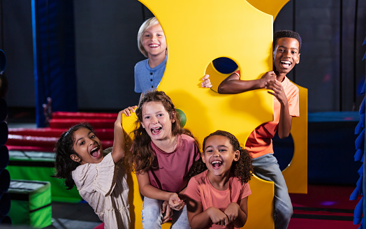 A multiracial group of five children having fun at an indoor amusement center, posing together around a yellow panel that looks like swiss cheese. They range in age from 7 years old to 10.