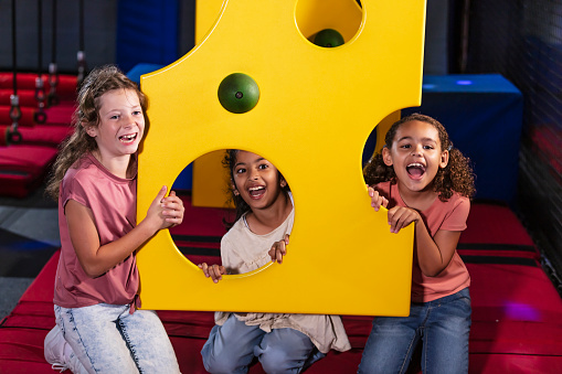 Three multiracial girls having fun at an indoor entertainment center, playing on a climbing wall that looks like a piece of swiss cheese. The African-American and Caucasian girl on the right is 7 years old. Her friends are 9 years old.