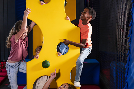 A multiracial group of five children having fun at an indoor amusement center, on a climbing wall that looks like swiss cheese. They range in age from 7 years old to 10.