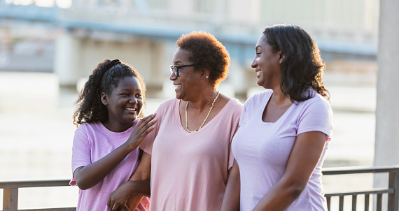 A multi-generation African-American family having fun walking together on a city waterfront. The three generations of women are smiling, holding hands. The senior grandmother is in the middle between her teenage granddaughter and her adult daughter.
