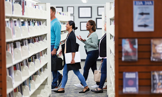 A multiracial group of five adult students walking together through a library past rows of bookshelves. The youngest, an Hispanic and Pacific Islander woman in her 20s, is carrying a laptop. Her friends are in their 40s and 50s.