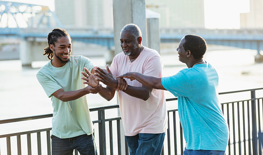 A multi-generation African-American family spending time together on a city waterfront. The three generations of men are walking side by side with the grandfather, in his 70s, in the middle. The grandson is a young man in his 20s. They are conversing, smiling, and interacting.