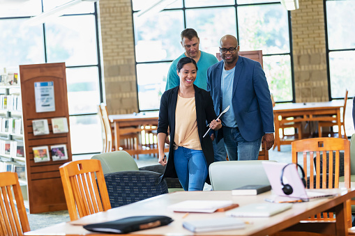 A multiracial group of adult students in the library, walking toward a table to work on a project together. The woman is in her 20s, Hispanic and Pacific Islander. The men are in their 40s and 50s.